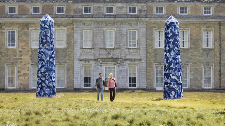 Two visitors walking past giant blue and white ceramic monoliths from the Expressions in Blue exhibition, in Petworth House's parkland with house in background.